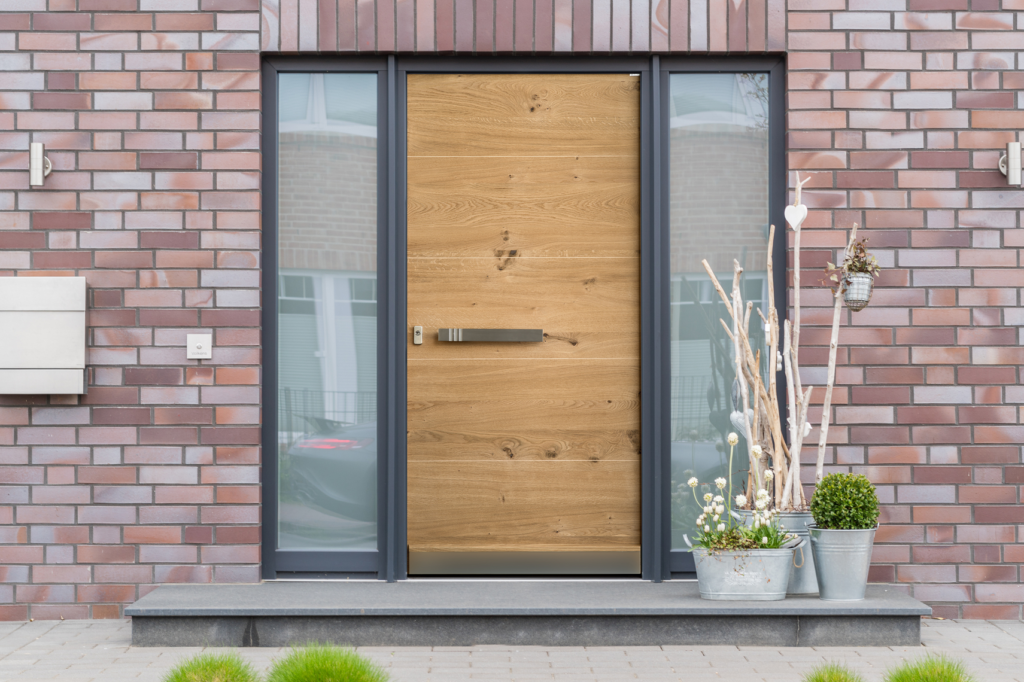 Moderner Hauseingang mit heller Holzhaustür, seitlichen Glasflächen und dekorativen Pflanzgefäßen vor einer roten Klinkerfassade.