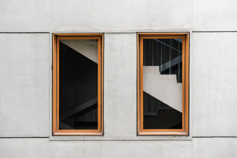 Zwei rechteckige Fenster mit Holzrahmen in einer grauen Betonfassade, durch die ein Treppenhaus sichtbar ist.