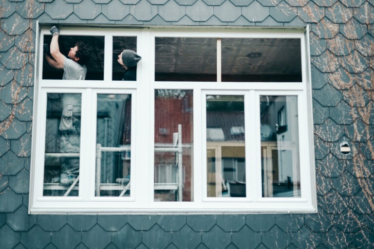 Zwei Handwerker installieren ein großes weißes Kunststofffenster an einer Hausfassade mit dunkler Schieferverkleidung.